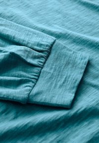 Dettaglio in tessuto turchese con polsino a manica arricciato, texture liscia e leggere variazioni naturali nel colore e nel motivo della tessitura.