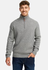 Ung mand smilende, iført en grå ribbet halv-zip sweater og mørkeblå jeans, med den ene hånd i lommen og lys baggrund.