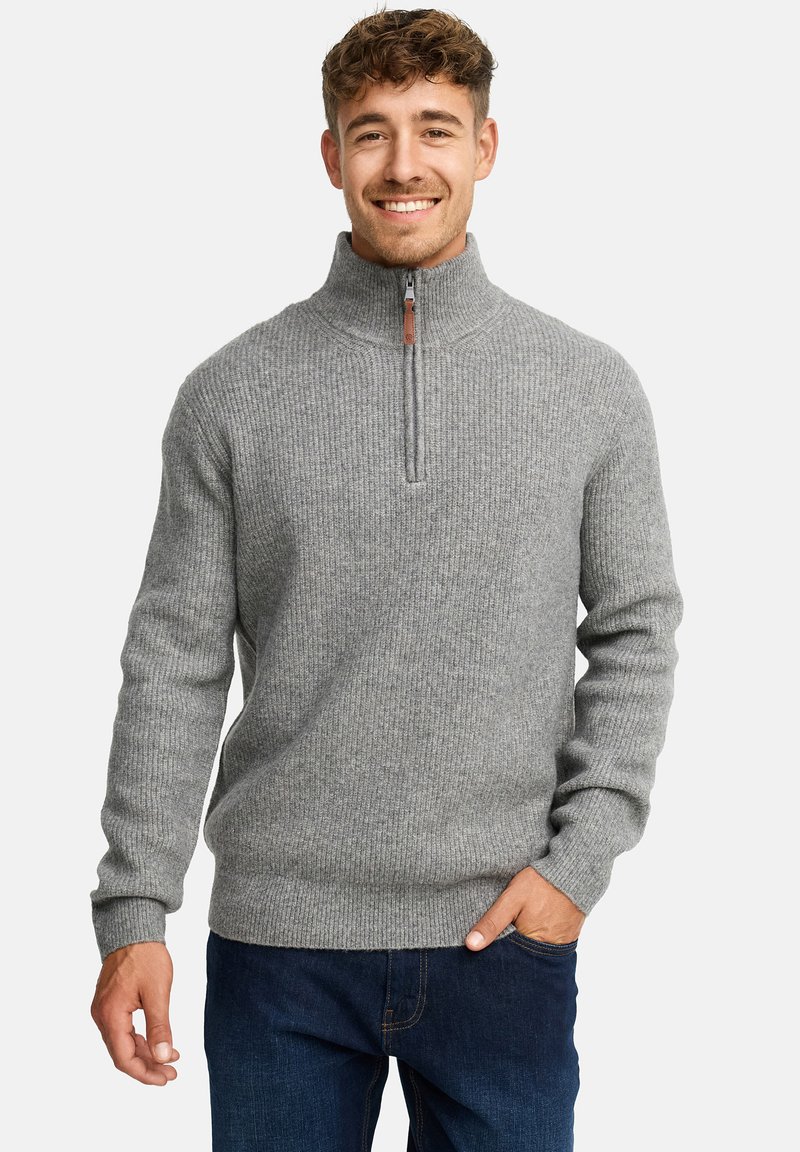 Ung mand smilende, iført en grå ribbet halv-zip sweater og mørkeblå jeans, med den ene hånd i lommen og lys baggrund.