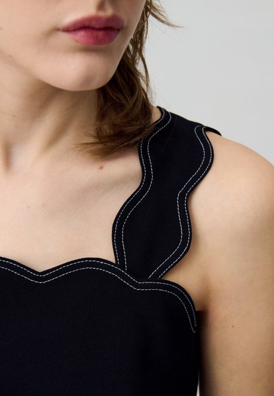 Haut noir avec décolleté festonné et accents de couture blanche. Texture lisse et design ajusté, mettant en valeur les épaules et la clavicule.