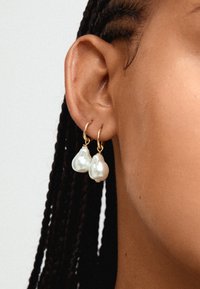 Brincos de argola dourados com duas pérolas irregulares e off-white suspensas em cada argola. As pérolas têm uma superfície lisa e refletora.