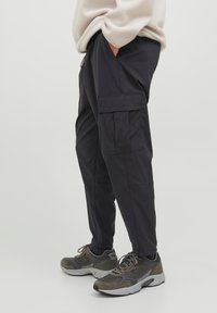 Pantaloni cargo neri con molteplici tasche, vita regolabile e design a gamba affusolata, abbinati a sneakers grigie. Tessuto liscio e leggero.