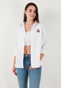 White button-up shirt made of cotton with a relaxed fit, featuring a bear embroidery on the left chest and rolled-up sleeves.