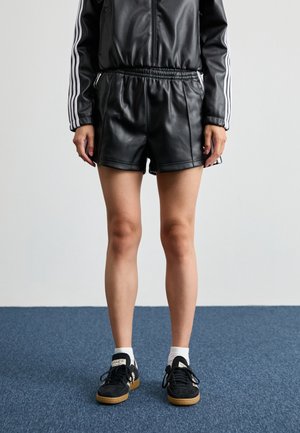Persoon draagt zwarte leren shorts, bijpassend jasje met witte strepen op de mouwen, witte sokken en zwarte sneakers staand op een blauw tapijt.