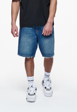 Shorts en denim bleu avec un ourlet effiloché, affichant une coupe décontractée. Associés à des baskets blanches et des chaussettes noir et blanc.