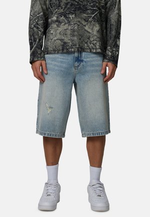 Jasnoniebieskie szorty jeansowe z postrzępionymi detalami i luźnym krojem. W zestawie z czarną koszulą z długim rękawem i białymi sneakersami.