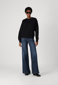 Felpa nera con colletto a girocollo, abbinata a jeans blu a gamba larga. Modella con occhiali da sole neri, in piedi su uno sfondo neutro.