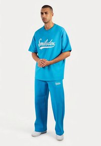 Conjunto azul de dos piezas que incluye una camiseta de corte holgado y pantalones a juego, ambos con el logo "Smilodox" en texto blanco. Textura de tela lisa.