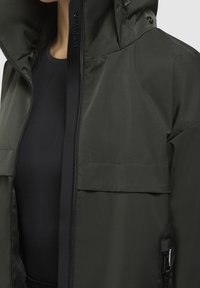 Mörkgrön vattentät jacka med dragkedja fram, stor huva och två sidofickor; slät yta med minimalistisk design.