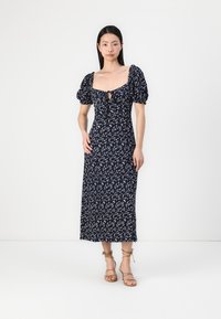 Vestido midi florido azul-marinho com mangas bufantes e detalhe em buraco. O tecido tem uma textura suave com pequenos padrões florais brancos. Combinado com sandálias com tiras.