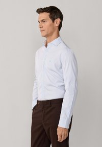 Muž v svetlom modrom pruhovanom košeli so zapínaním na gombíky, zastrčený v tmavohnedých nohaviciach, stojaci bokom na jednoduchom pozadí.