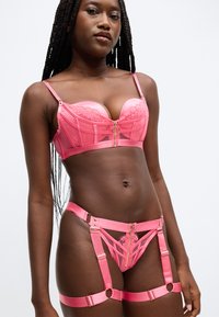 Ensemble de lingerie rose comprenant un soutien-gorge en satin et dentelle, une fermeture éclair avant, et une jarretelle assortie avec des bretelles réglables et des accents dorés.