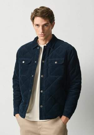 Man wearing a navy blue quilted jacket over a white shirt and beige pants, standing with hands in pockets against a plain background.