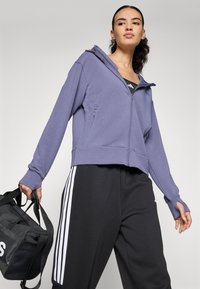 Lila, verkürzter Hoodie mit Reißverschluss, langen Ärmeln und seitlichen Taschen; getragen mit schwarzen Hosen mit weißen Seitenstreifen und einer schwarzen Tasche.