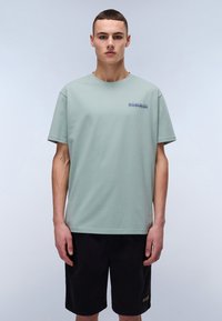 Světlounce zelené bavlněné tričko s kulatým výstřihem a krátkými rukávy, na prsou se modrou vyšívanou nášivkou. Model má na sobě černé kraťasy.