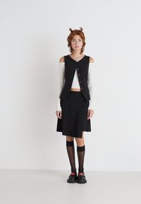 Black vest with buttons over a white crop top. Black pleated skirt, knee-high socks, and chunky black shoes. Simple, layered outfit.