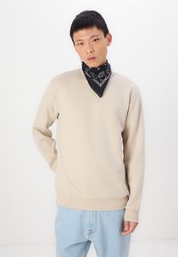 Beige tröja med rund halsringning i mjukt material, med ribbade muddar och fåll. Bärs med en svart bandana med vita mönster. Ljust blå jeans.