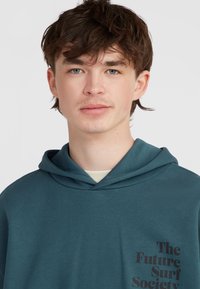 Turkizna kapuca s velikim prednjim džepom, podesivim trakama i crnim grafičkim otiskom na lijevoj strani prsnog područja. Glatka tekstura tkanine.