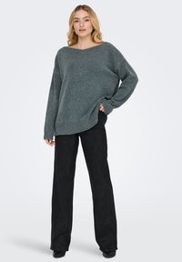 Prevelik sivi pulover z širokim ovratnikom in rebrastimi robovi, kombiniran s črnimi širokimi hlačami, izdelan iz mehke, teksturirane tkanine.