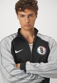 Αθλητική ζακέτα Nike με μαύρο σώμα, γκρι μανίκια, κλείσιμο με φερμουάρ μπροστά και λογότυπο με θέμα το μπάσκετ στο στήθος.