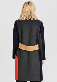 Abrigo de lana gris con un cinturón mostaza, con mangas navy y un acento naranja intenso en el lado inferior izquierdo. Diseño sencillo, hasta la rodilla.