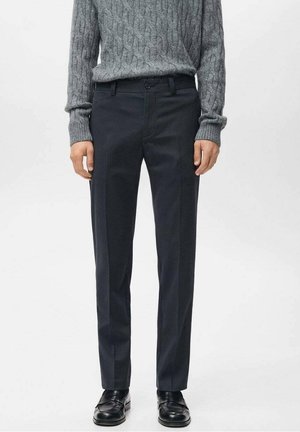 Mann trägt dunkle, maßgeschneiderte Hose, schwarze Loafers und einen grauen Zopfpullover vor neutralem Hintergrund.