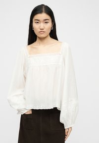 Jeune femme aux longs cheveux noirs portant un chemisier blanc orné de dentelle avec des manches bouffantes et une jupe sombre, se tenant devant un fond uni.