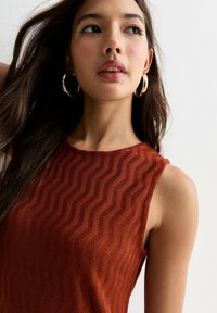 Top sans manches couleur rouille avec un motif texturé en vagues, encolure ronde et tissu lisse. Le mannequin porte des boucles d'oreilles créoles dorées et a les cheveux longs.