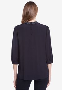 Blusa nera con maniche tre quarti, colletto rotondo con pois bianchi e chiusura con bottoni sul retro. Tessuto morbido e leggero.