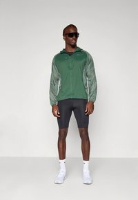 Green lightweight jacket with a zip front and gray sleeves, paired with black shorts. Model wears sunglasses and white sneakers.