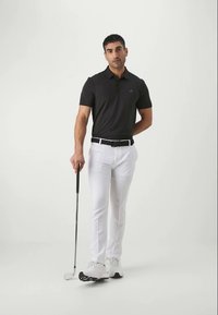 Muškarac u crnoj polo majici i bijelim hlačama drži golf palicu, stojeći uz jednostavnu bijelu pozadinu.