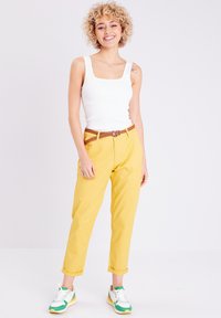 Pantalons en coton jaunes avec des poignets retournés, associés à un débardeur blanc à côtes et des baskets à accents verts. Ceinture tressée marron en détail.