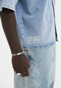 Lyseblå denimskjorte med korteærmer og hvidt logo; akkompagneret af matchende denimbukser med et diskret mønster; sølv smykkedetaljer.