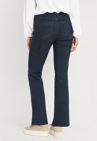 Donkerblauwe flared jeans met een tailleband, twee achterzakken en een gladde textuur. G gedragen met beige schoenen.