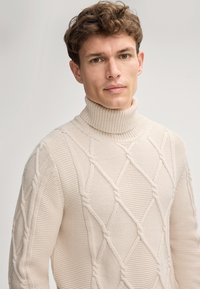 Beige Rollkragenpullover mit einem strukturierten Zopfmuster, das diamantförmige Formen sowie eine gerippte Kragen- und Ärmelabschlüsse aufweist.