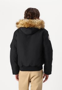 Black bomber jacket with a fur-lined hood, ribbed cuffs, and a logo patch on the sleeve. Smooth texture and relaxed fit.