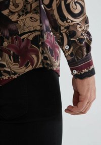 Chemise à manches longues avec un imprimé floral et animal complexe dans des couleurs sombres, avec un poignet et des détails de bouton en bordeaux contrastants.