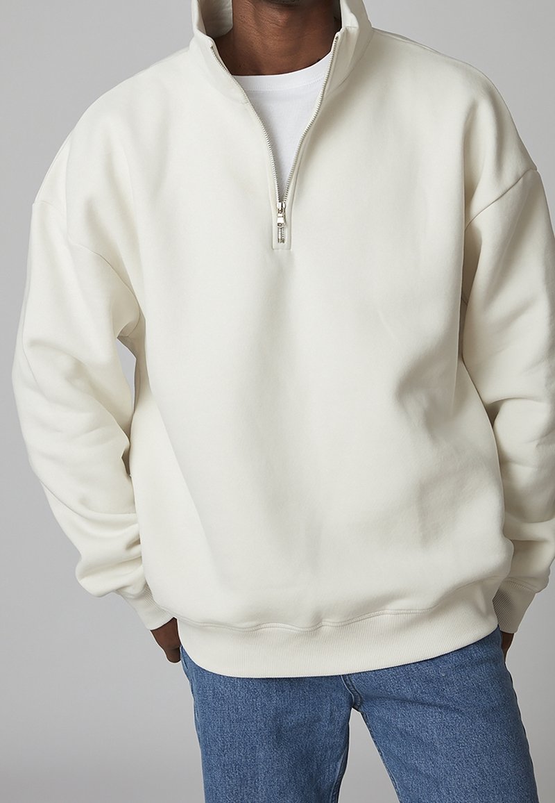 Sudadera de forro polar color crema con cuello alto y cremallera media. Tiene un corte relajado y puños y dobladillo acanalados. Combinada con jeans azules.