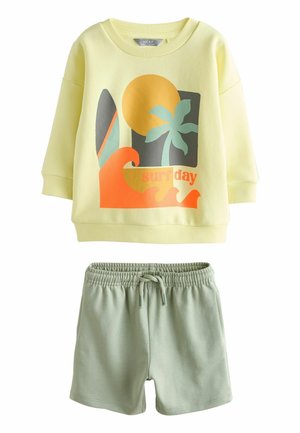 Jasnożółta bluza z grafiką o tematyce surfingowej i długimi rękawami, zestawiona z zielonymi szortami na elastycznej gumce w pasie. Miękka tkanina, casualowy fason.