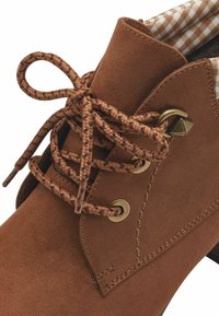 Brown suede ankle boot featuring a checkered lining, round toe, and braided lace with gold-tone hardware accents.