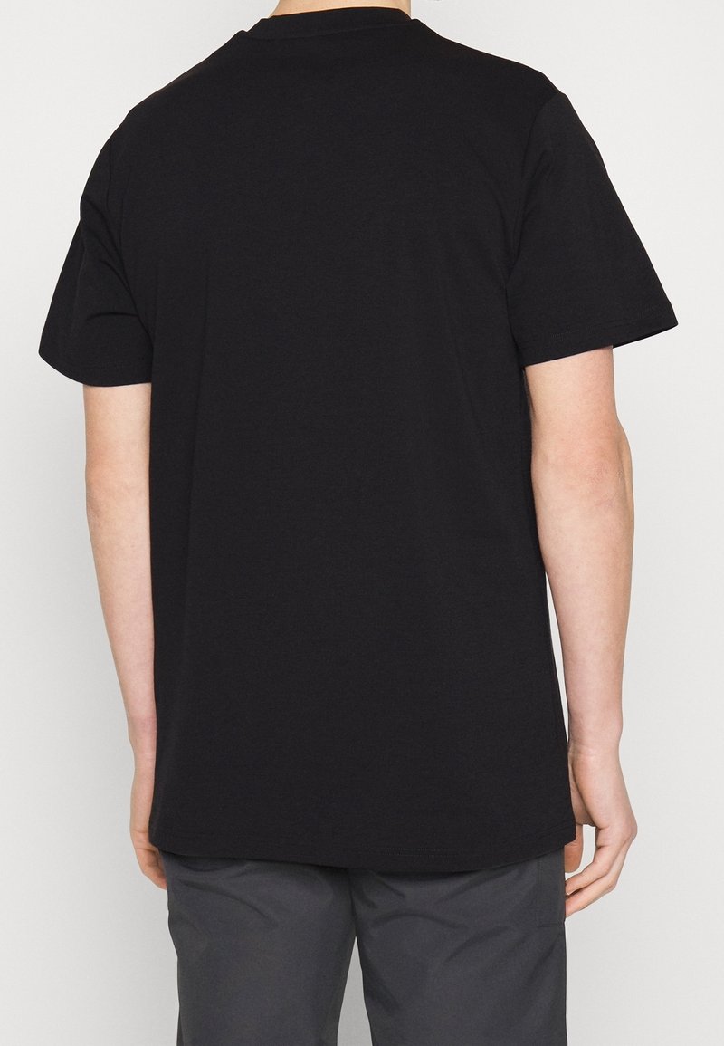 Personne portant un t-shirt noir uni à manches courtes vue de dos, associée à un pantalon gris foncé sur fond blanc uni.