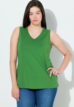 Junge Frau mit langen, dunklen Haaren, die ein ärmelloses grünes Oberteil und blaue Jeans trägt und mit einer Hand in der Hüfte vor einem weißen Hintergrund steht.