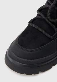 Czarne zamszowe sneakersy z teksturowaną gumową podeszwą, z ciemnymi sznurówkami i wyściełanym kołnierzem. Gładkie akcenty kontrastują z szorstkim wzorem podeszwy.