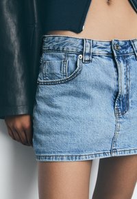 Lichtblauwe denim minirok met bruine stiksels, met een knoopsluiting aan de voorkant, riemlussen en twee zijzakken.