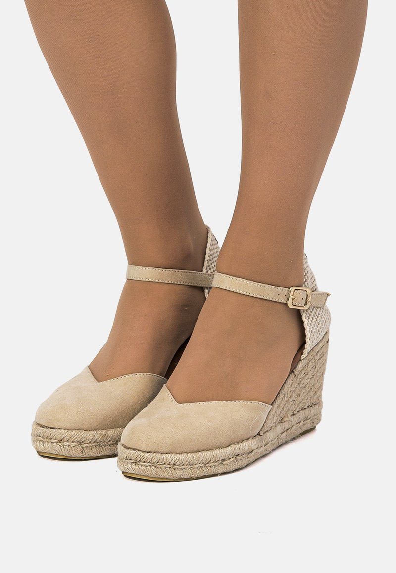 Sandalias cuña tipo esparto beige con parte superior de ante, diseño de punta en forma de V y tacón tejido. Correa ajustable en el tobillo con detalle de hebilla metálica.