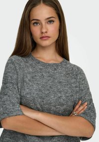Sivi pleteni pulover s kratkim rukavima i rebrastim ovratnikom. Mekana tekstura s malo labavim krojem. Model pozira s prekriženim rukama.