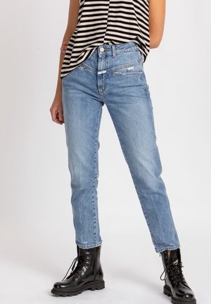 Henkilö, jolla on vaaleansiniset korkeavyötäröiset farkut, mustat nauhalliset sotilasaappaat ja mustavalkoraidallinen hihaton paita.