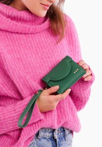 Green textured wallet with a curved flap, three decorative studs, and a wrist strap, held against a pink sweater background.
