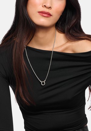 Donna con lunghi capelli scuri che indossa un top nero scollato e una collana d'argento con un ciondolo circolare.