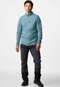 Giacca in pile di colore azzurro chiaro con colletto alto, zip frontale e tasca sul petto con zip, abbinata a pantaloni cargo neri con tasche con zip.